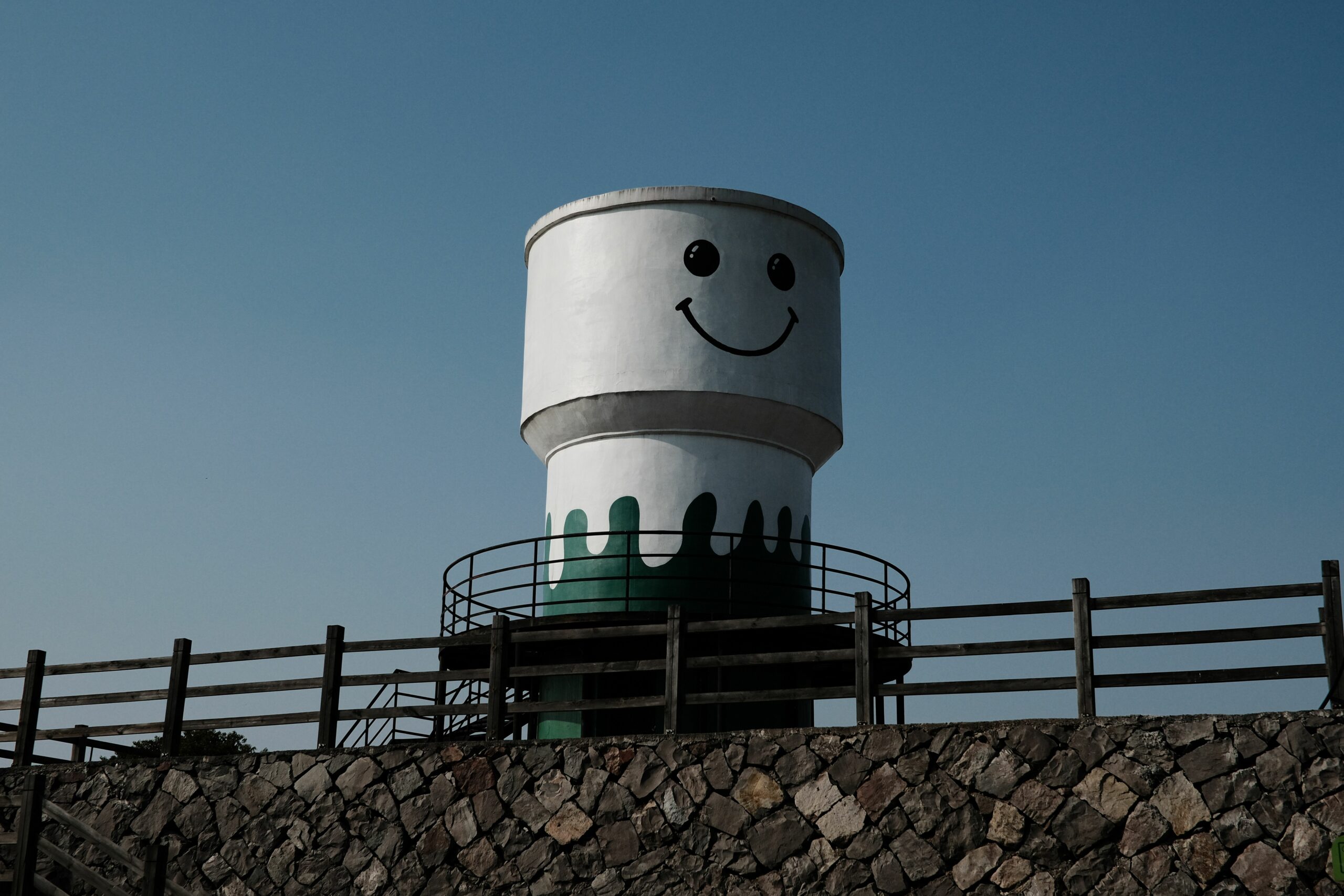 Artistic water tower with a smiley face under a clear blue sky, surrounded by a fence and stone wall.